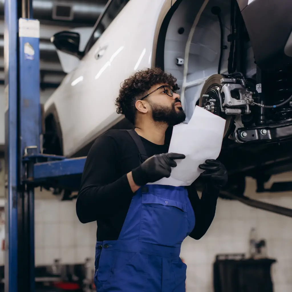 A mechanic holds a document while inspecting beneath a lifted car.