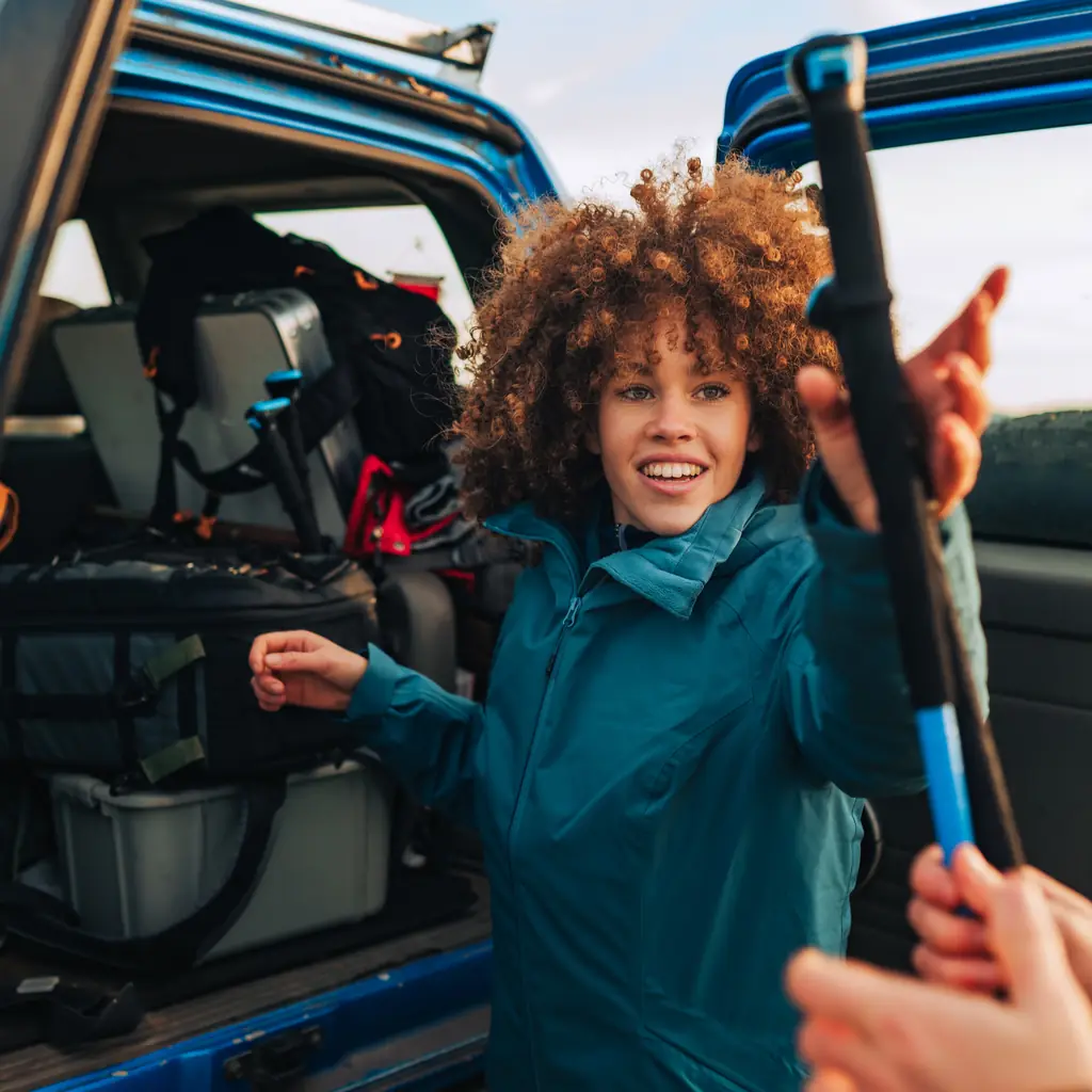 A hiker retrieves trekking poles beside an open vehicle.