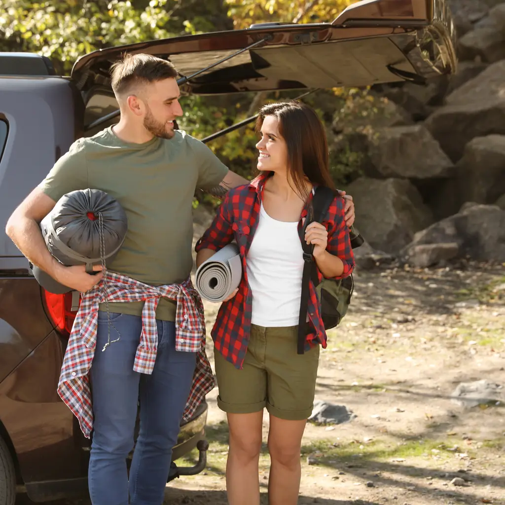 Two campers carry a rolled mat and sleeping bag beside an open vehicle.