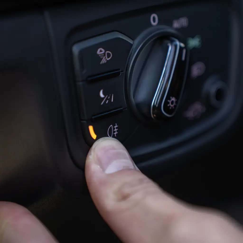 A finger pressing a rear fog-light button beside the headlight selector.
