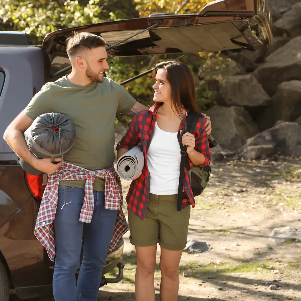 Two campers carry a rolled mat and sleeping bag beside an open vehicle.