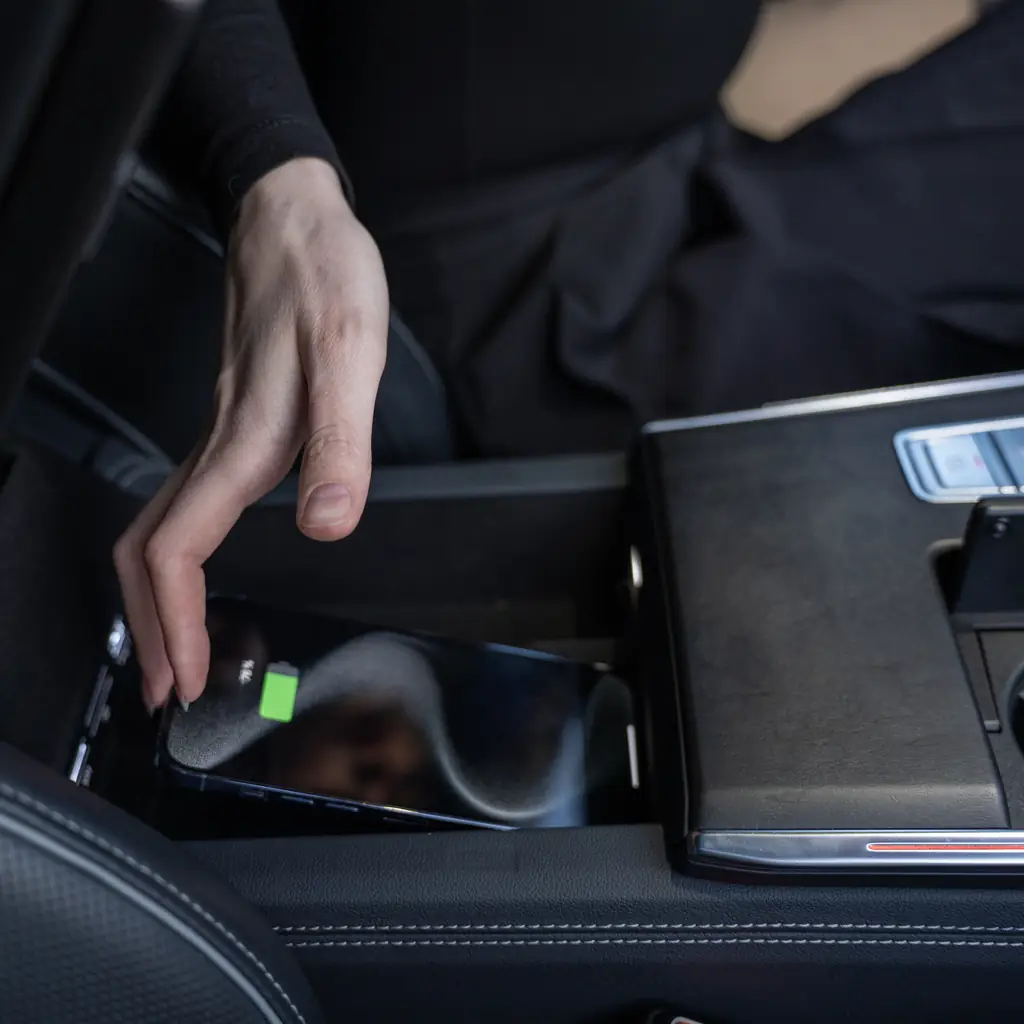 A hand lifts a smartphone from a wireless charging pad in a center console.