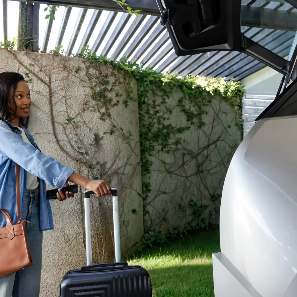 A traveler checks a phone while moving a suitcase beside an open vehicle hatch.